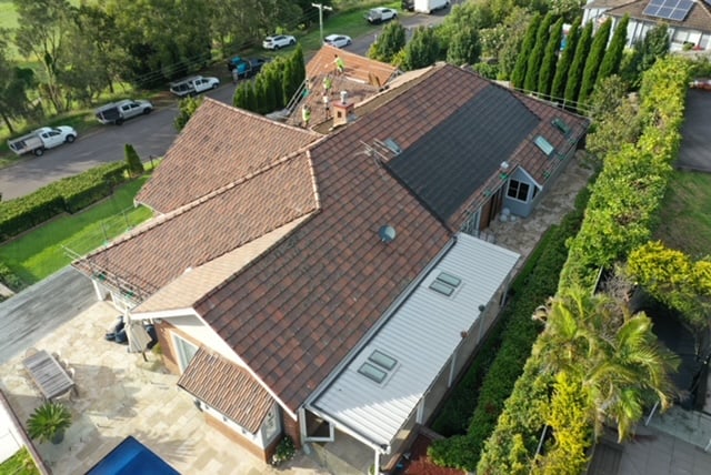 High-angle aerial view of a large residential home undergoing roof restoration with tradespeople working near the ridge.